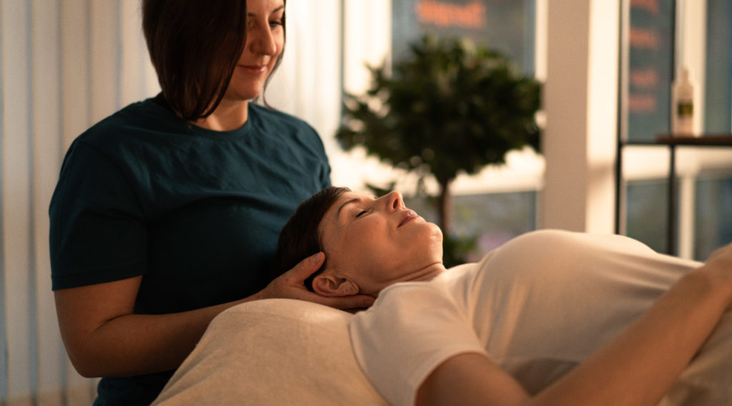 Eine Therapeutin stützt den Kopf einer entspannt auf einer Behandlungsliege liegenden Frau in einem hellen Behandlungsraum.