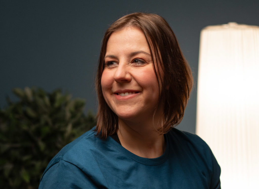 Eine lächelnde Therapeutin in blauem Oberteil sitzt in einem freundlich beleuchteten Raum und blickt zur Seite.