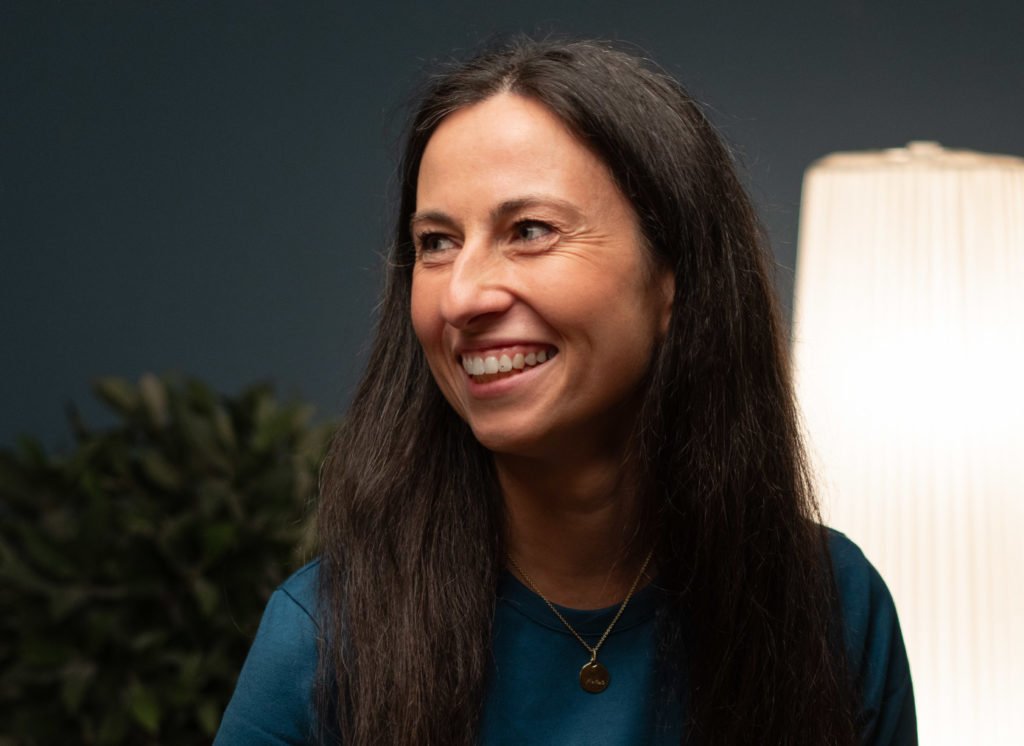 Eine lächelnde Therapeutin in blauem Oberteil sitzt in einem freundlich beleuchteten Raum und blickt zur Seite.