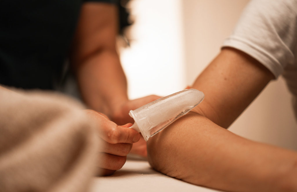 Eine Person hält eine Stange Eis an den Knöchel einer liegenden Person, während der Fuß auf einer Behandlungsliege ruht.