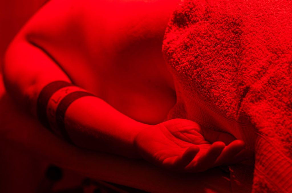 Nahaufnahme einer liegenden Person auf einer Behandlungsliege; eine Hand ruht geöffnet neben dem Körper, der Oberkörper ist mit einem Handtuch bedeckt und in rotes Licht getaucht.