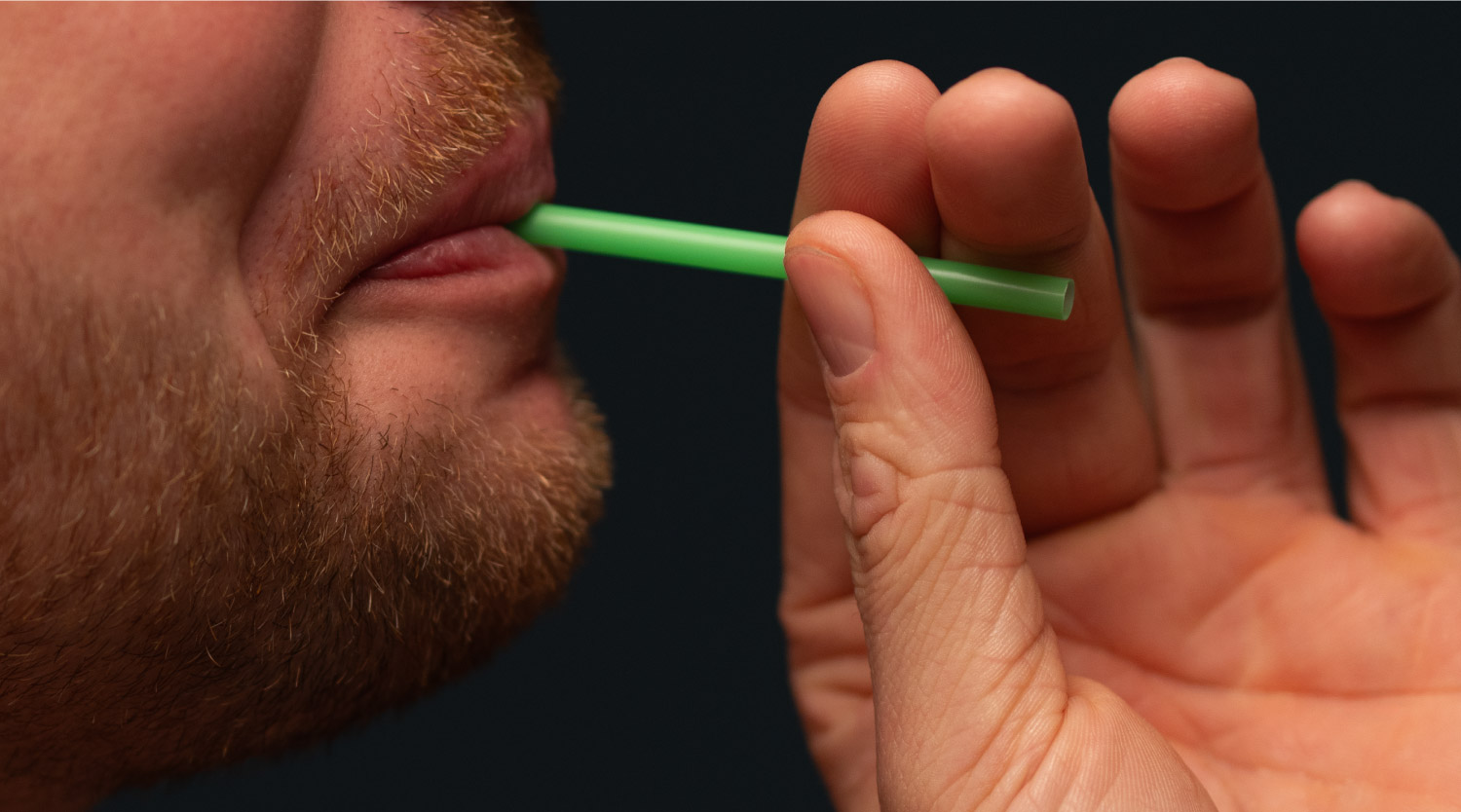 Nahaufnahme eines bärtigen Gesichts, das einen grünen Strohhalm zwischen den Lippen hält; eine Hand führt den Halm zum Mund.