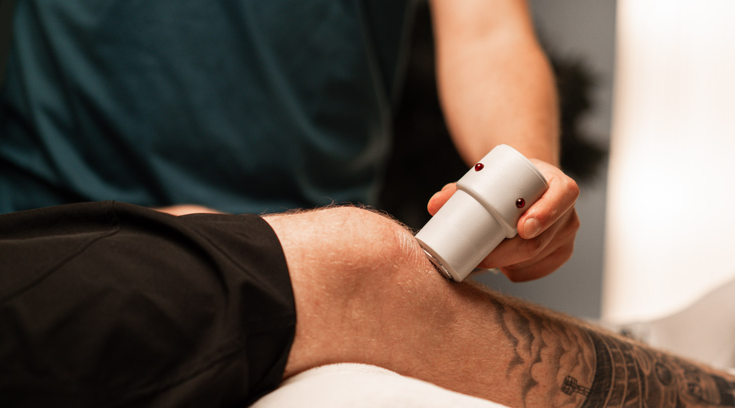 Eine Person hält ein weißes Therapiegerät an das Knie einer liegenden Person; der Unterschenkel ist teilweise tätowiert.