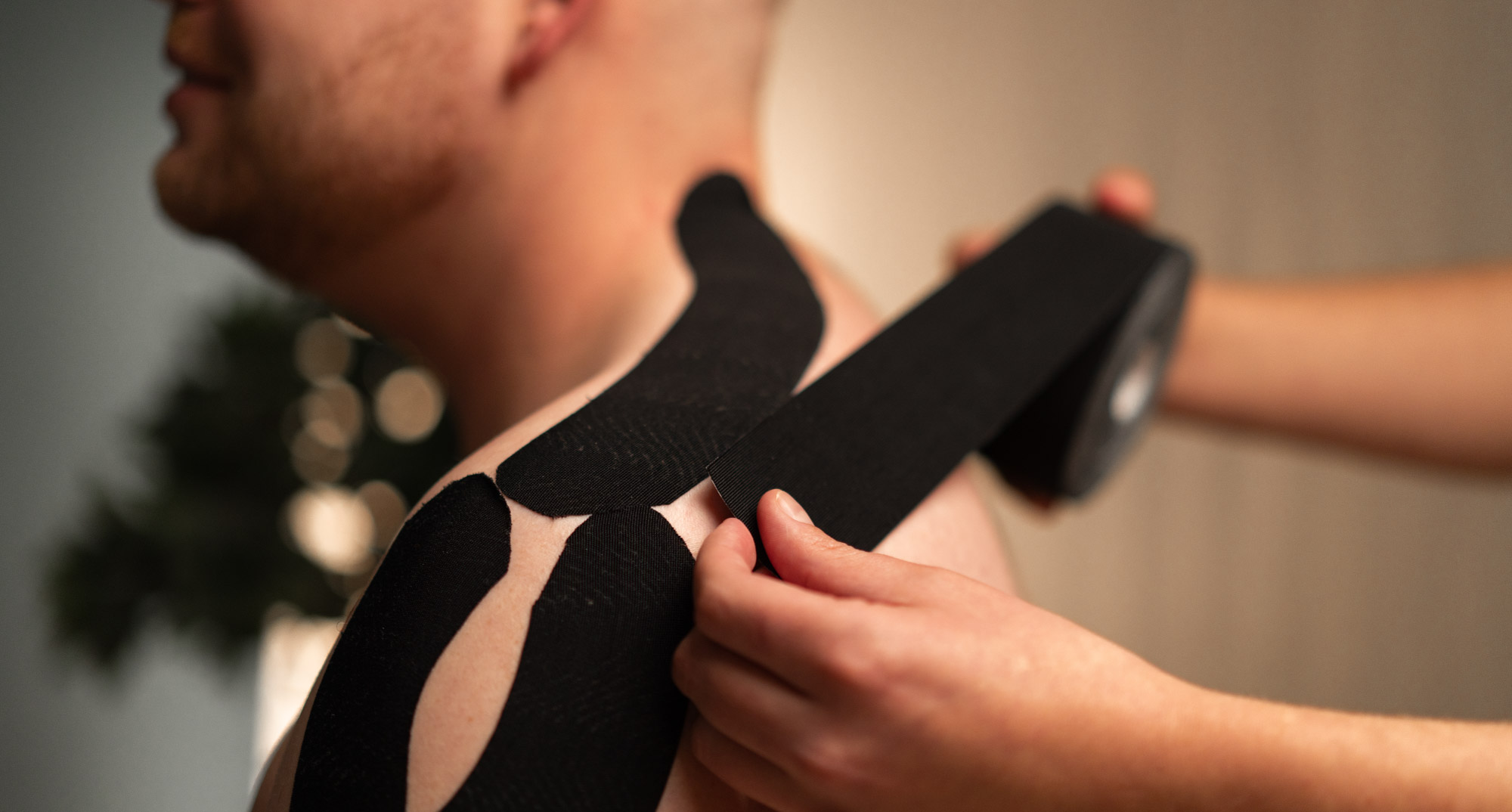 Eine Hand klebt schwarzes Kinesiotape auf Schulter und Nacken eines sitzenden Mannes.