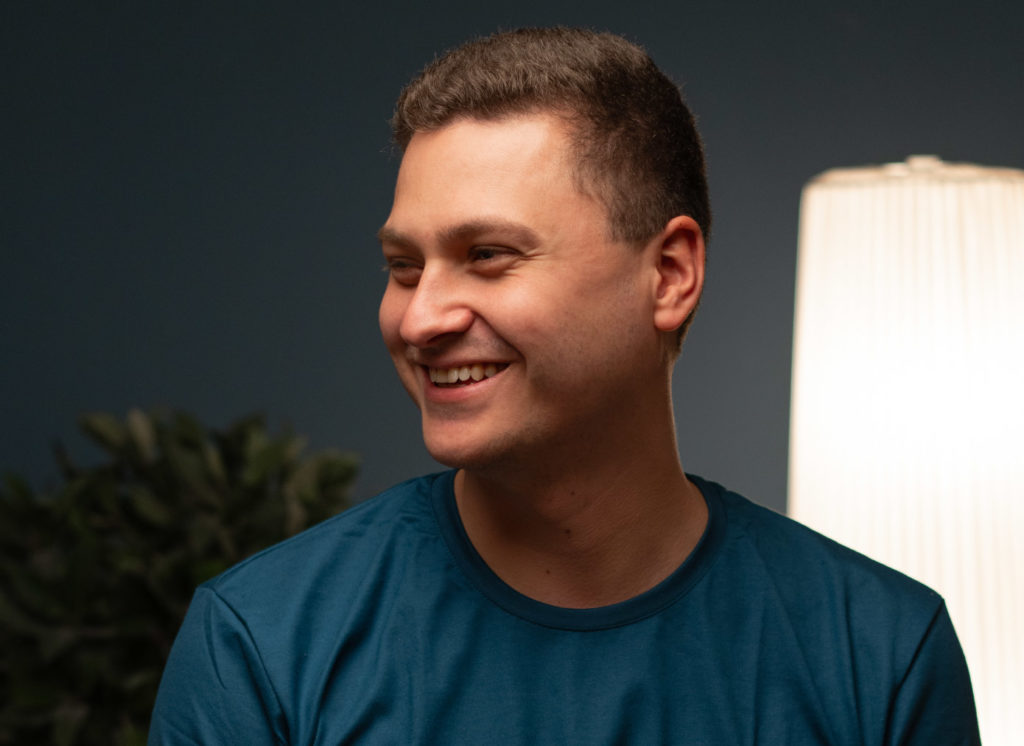 Ein lächelnder Therapeut in blauem Oberteil sitzt in einem freundlich beleuchteten Raum und blickt zur Seite.