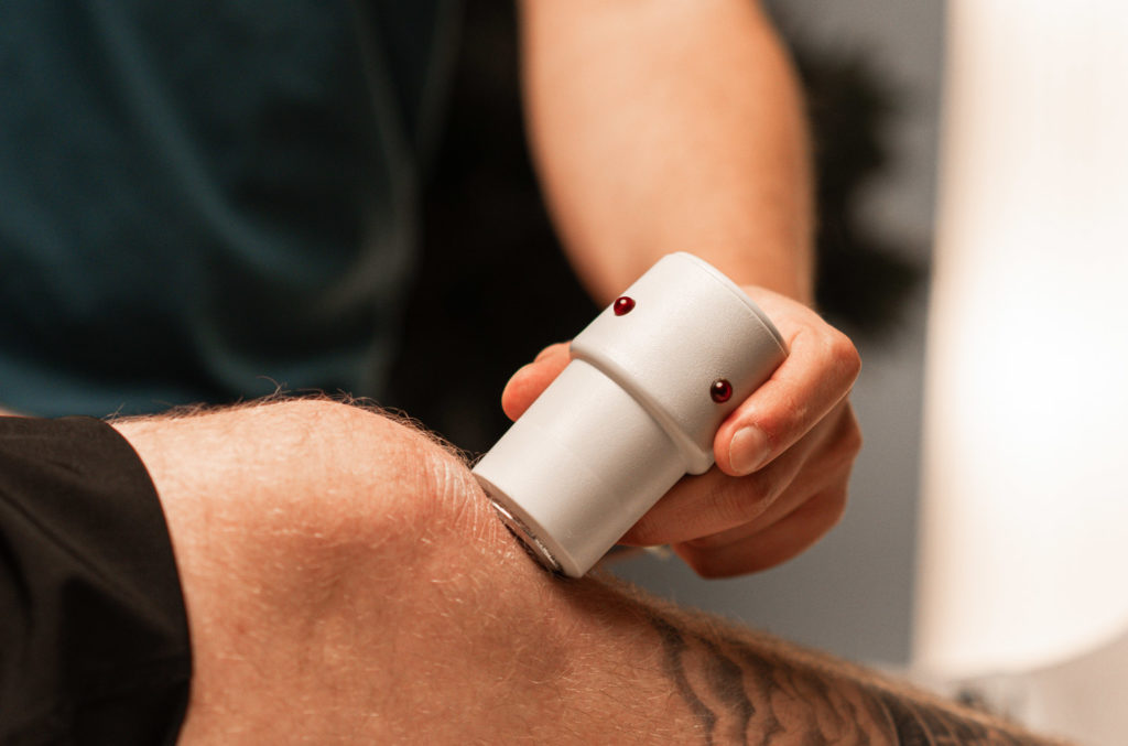 Eine Person hält ein weißes Therapiegerät an das Knie einer liegenden Person; der Unterschenkel ist teilweise tätowiert.
